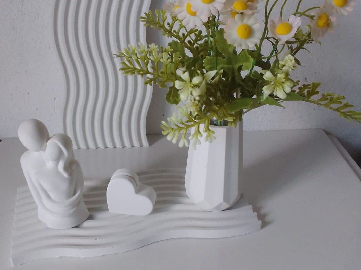 Figura de pareja, corazón y florero geométrico sobre una bandeja ondulada de cemento blanco